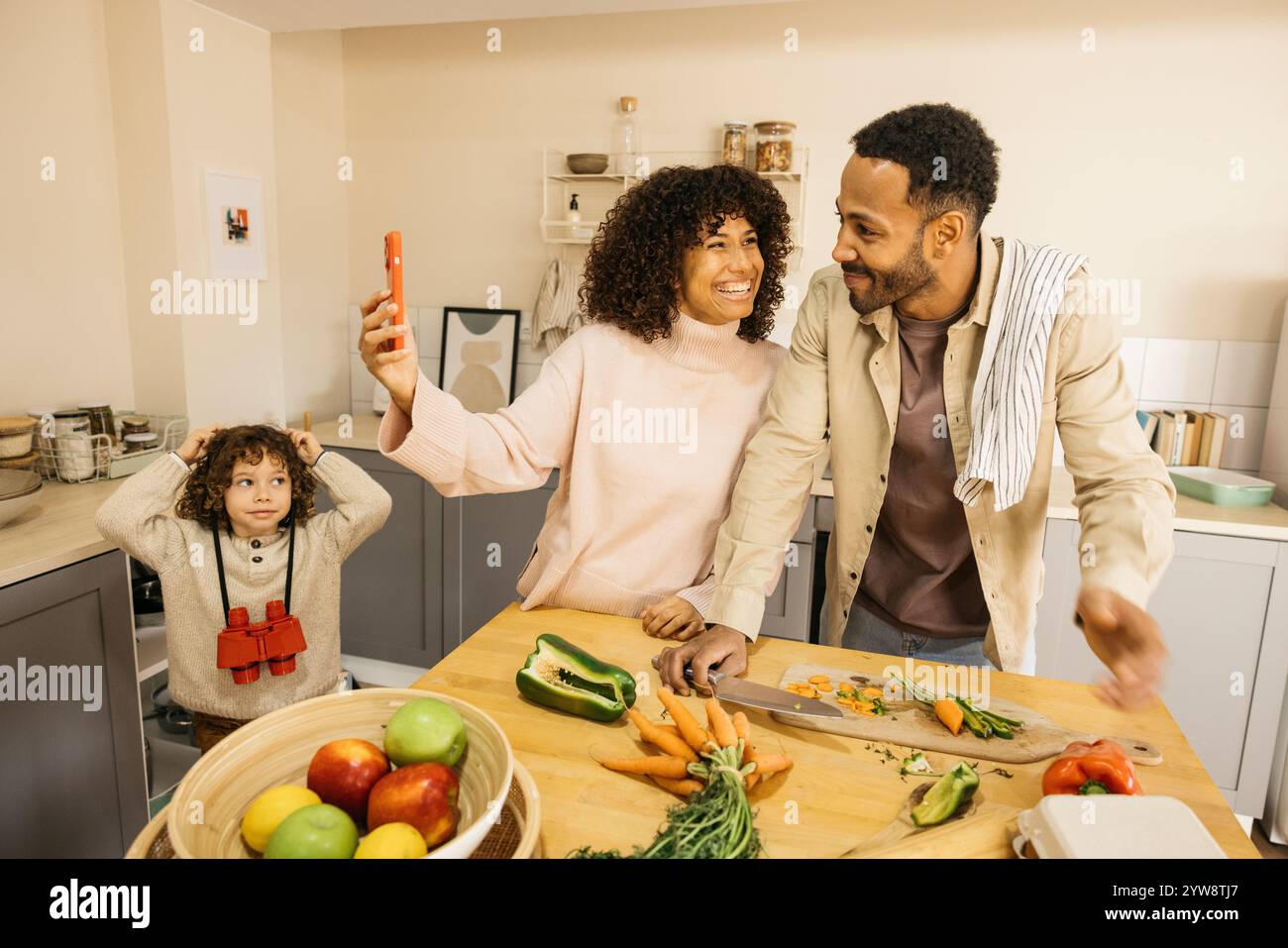 Una famiglia gioiosa che prepara un pasto mentre scatta foto in cucina. La scena cattura la felicità e un'atmosfera giocosa, con verdure fresche Foto Stock