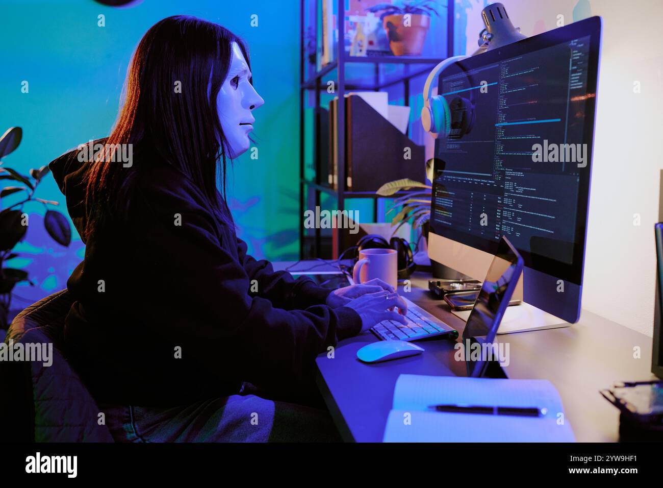 Immagine laterale di una hacker seduta sulla scrivania che guarda il monitor mentre scrive i codici sulla tastiera, indossa una maschera bianca e una felpa con cappuccio nera Foto Stock