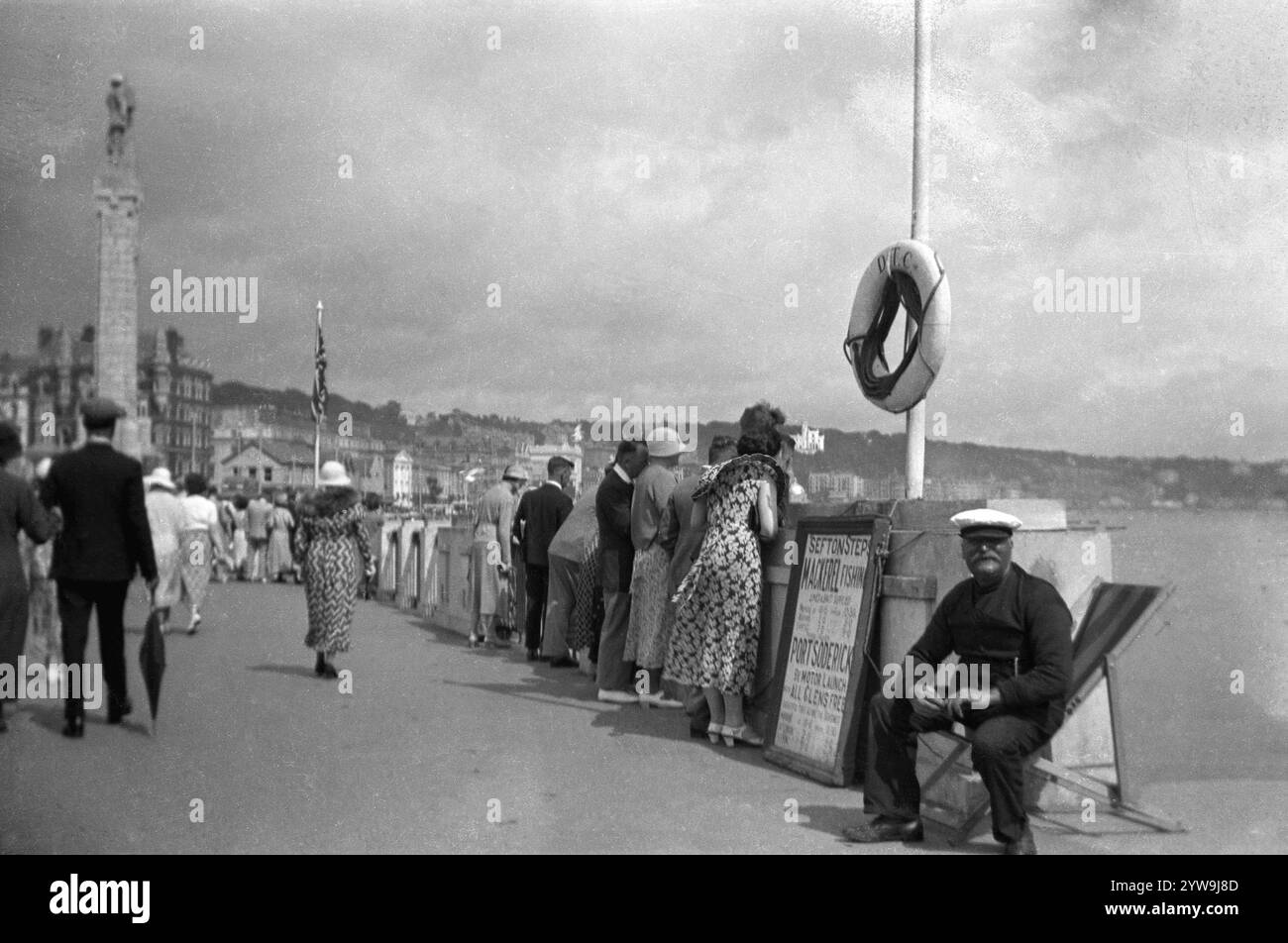 Anni '1930, storico, gente sul lungomare Douglas, Isola di Man, Inghilterra, Regno Unito. Fisherman in cap seduto su una sdraio accanto a un cartello con scritto Stefton Steps, pesca dello sgombro e gite di un giorno a Port Soderick con "All Glens Free". La spiaggia di Port Soderick è un'area tranquilla e tranquilla a pochi chilometri da Douglas. Foto Stock