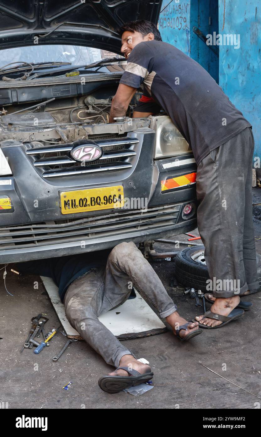 Due giovani meccanici indiani a Mumbai, India, che lavorano al motore di un'auto, uno dei quali lavora sotto il veicolo Foto Stock