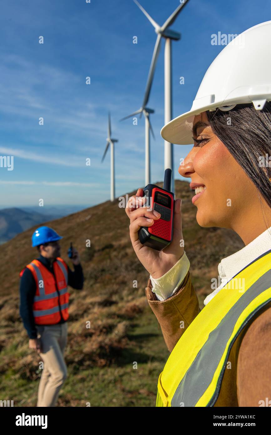 Gli ingegneri latini, donne e uomini, che indossano giubbotti di sicurezza e caschi, utilizzano walkie talkie per comunicare in un parco di turbine eoliche, lavorando su ener verde Foto Stock