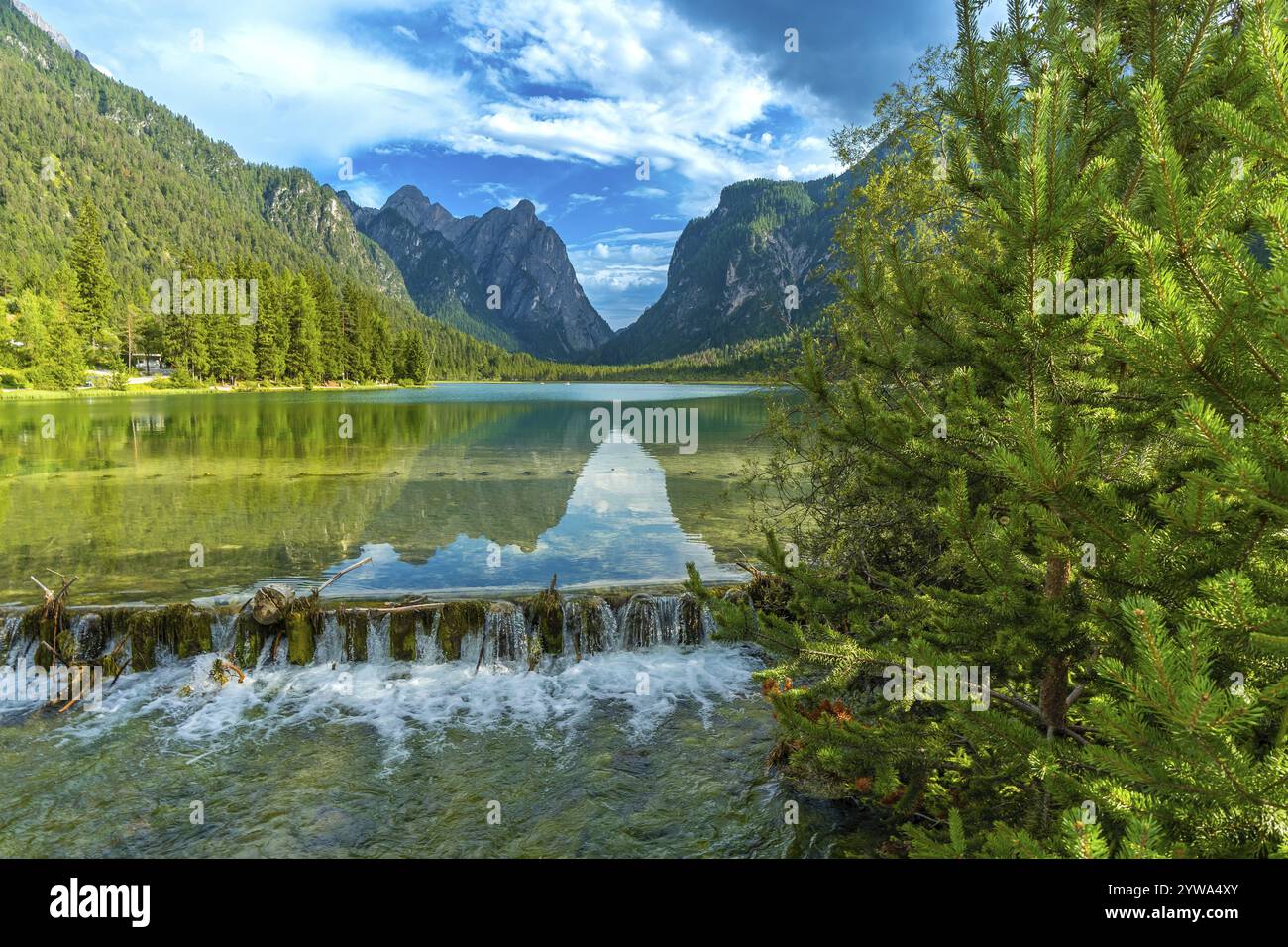 Acqua cristallina che si getta da una piccola cascata nel lago dobbiaco, riflettendo le maestose cime dolomitiche sotto il cielo estivo Foto Stock