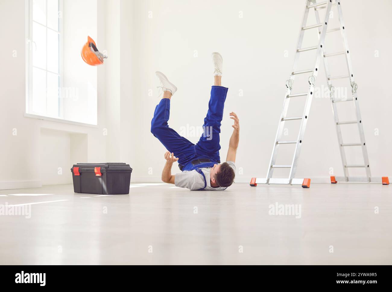 Giovane lavoratore in uniforme e casco di sicurezza caduto all'alba sul pavimento dalla scala Foto Stock