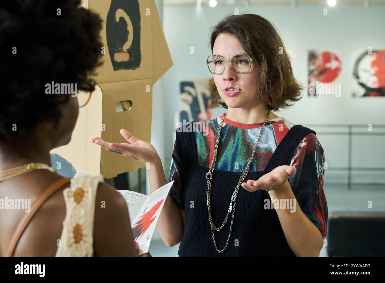Giovane donna bruna in occhiali da vista che spiega la sua opinione sulle opere d'arte rappresentate al visitatore afroamericano Foto Stock