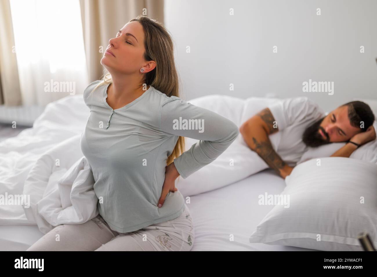 Una giovane coppia sta dormendo a letto. La donna si sta svegliando per lavoro. Ha mal di schiena. Foto Stock