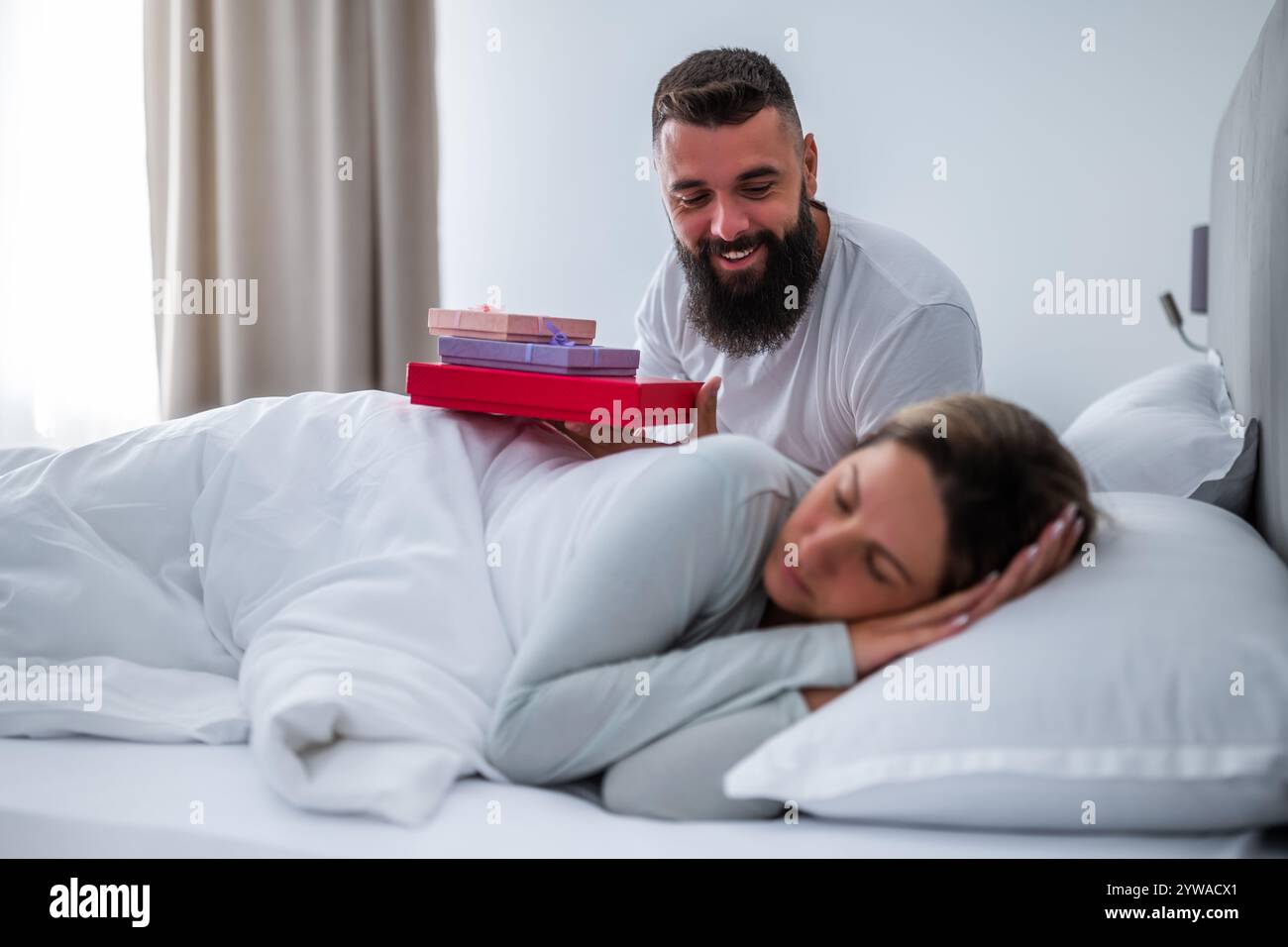 La giovane coppia è sdraiata a letto la mattina. L'uomo sta dando regali a sua moglie per il suo compleanno. Foto Stock