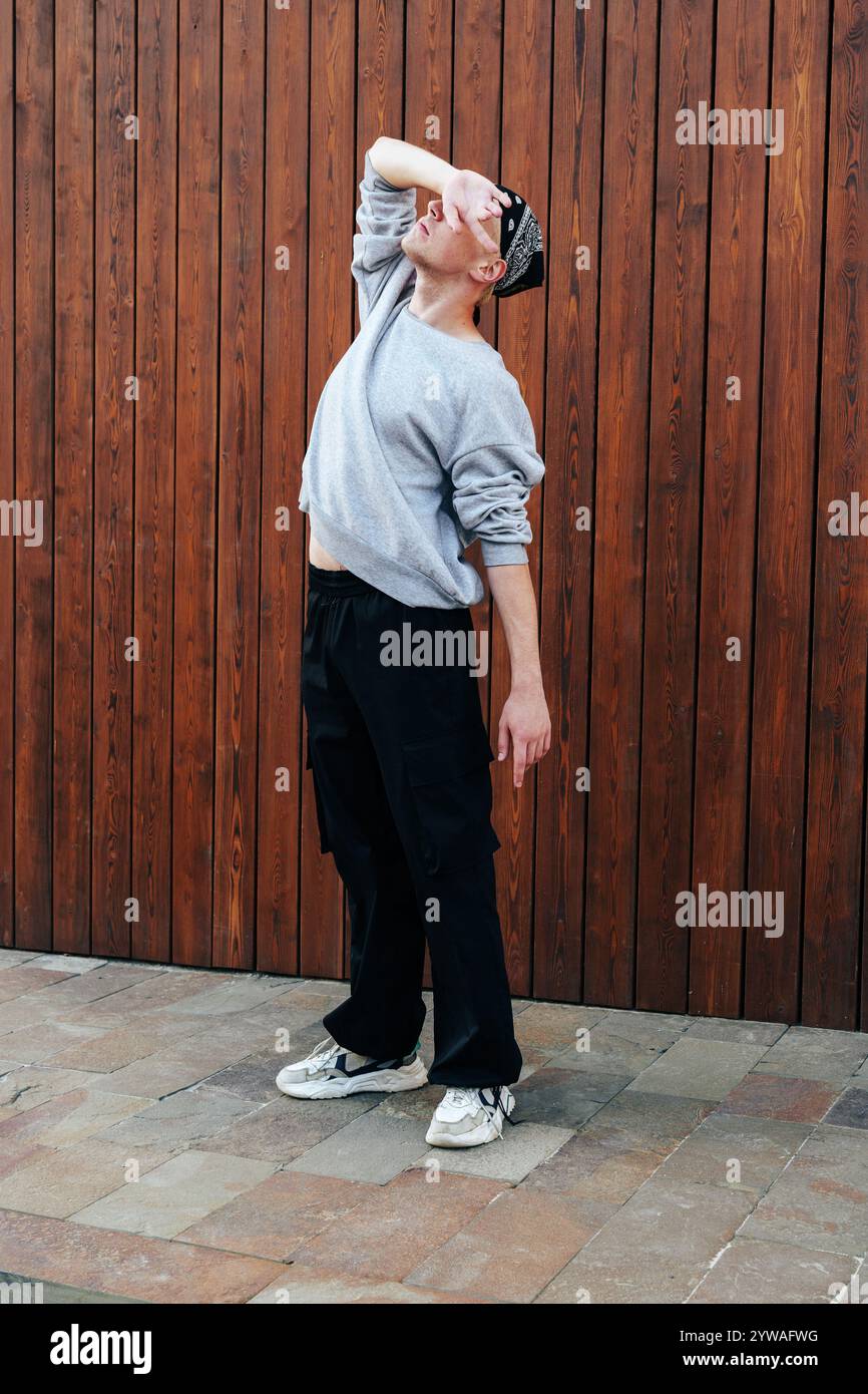 Una persona con una felpa grigia e pantaloni neri si mette in posa davanti a una parete di legno Foto Stock