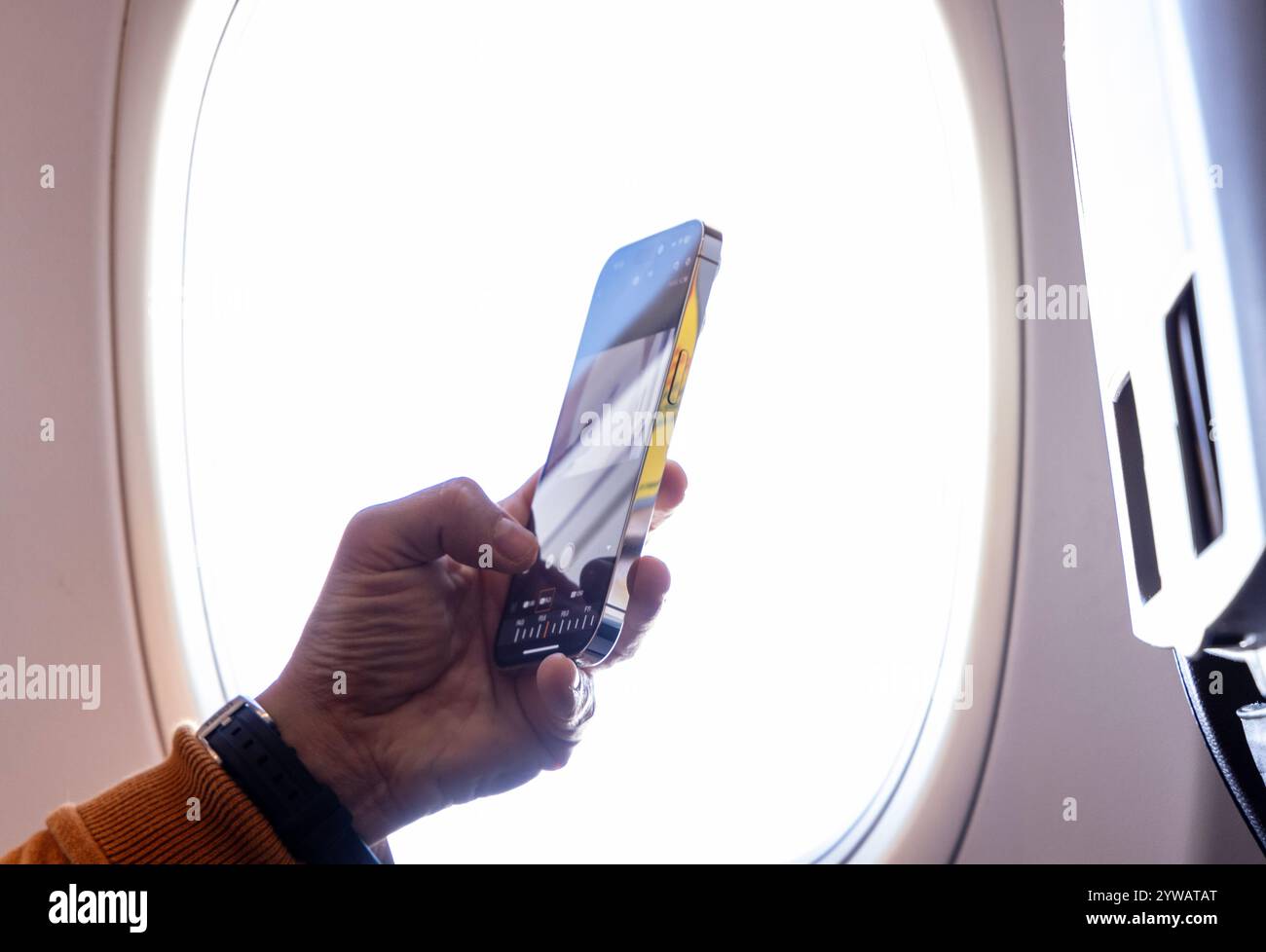 Primo piano di una mano che tiene uno smartphone vicino alla finestra di un aereo, catturando l'esperienza di viaggio e la connettività moderna durante un volo in un luminoso Foto Stock