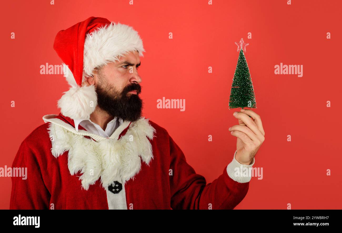 Buon Natale e felice anno nuovo. Babbo Natale ha in mano un piccolo albero di Natale. Ritratto di un uomo barbuto serio in abiti di Babbo Natale con piccolo d Foto Stock