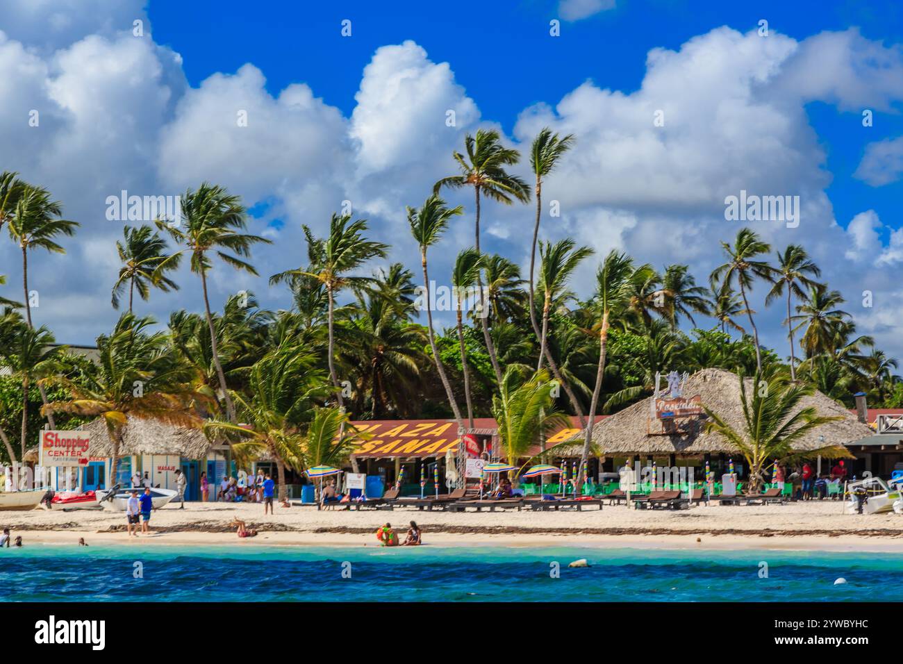 Una spiaggia con palme e alcuni edifici. La spiaggia è affollata di persone e ci sono alcune barche in acqua Foto Stock