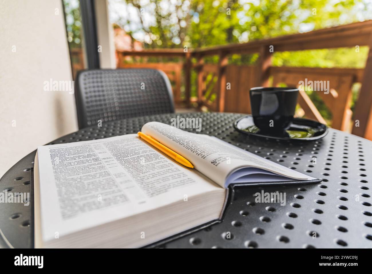 Apri la Sacra Bibbia con penna gialla adagiata su un tavolo da giardino in plastica con tazza da caffè nera in piedi sul balcone con recinzione in legno e dintorni verdi Foto Stock