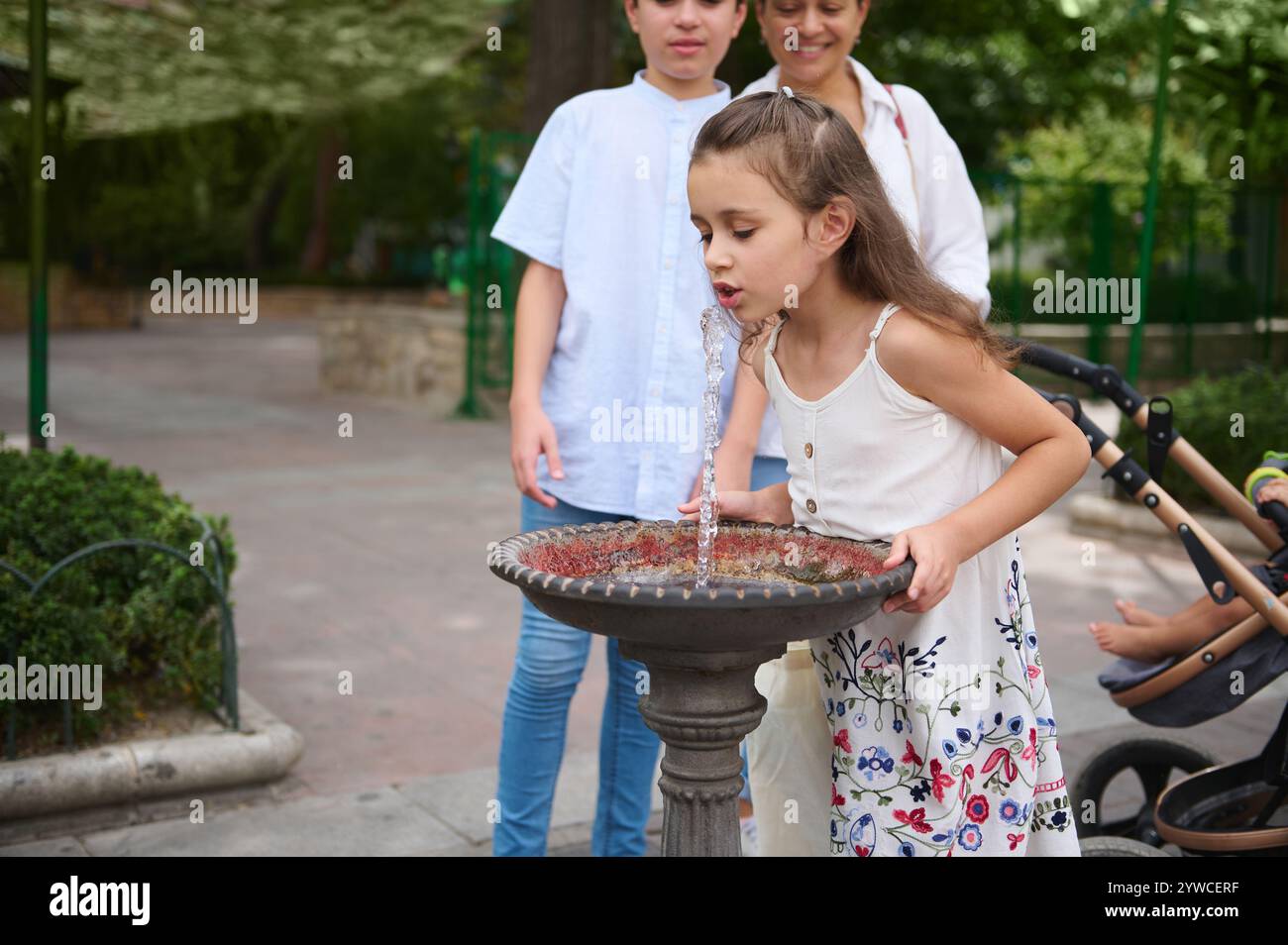 Una giovane ragazza si gode un drink rinfrescante da una fontana del parco con la sua famiglia nelle vicinanze. Foto Stock