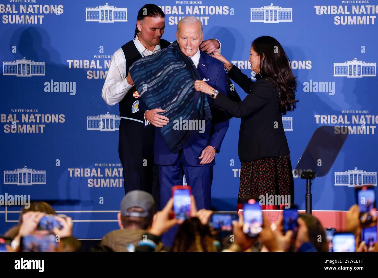 Il Segretario degli interni degli Stati Uniti Deb Haaland (R) e l'Assistente Segretario per gli affari indiani Bryan Newland (L) presentano al presidente degli Stati Uniti Joe Biden una coperta di ottava generazione proveniente da un'azienda di proprietà tribale ricamata con "Joe Biden Champion for Indian Country prima delle osservazioni al vertice delle Nazioni tribali della Casa Bianca del 2024 al Dipartimento degli interni di Washington, DC, USA, 09 dicembre 2024. L'amministrazione Biden-Harris ha dato priorità al rafforzamento delle relazioni nazione-nazione, onorando la fiducia e gli obblighi del trattato con le tribù riconosciute federalmente, e avanzando la sovranità tribale Foto Stock