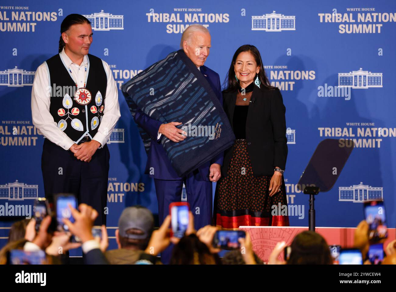Il Segretario degli interni degli Stati Uniti Deb Haaland (R) e l'Assistente Segretario per gli affari indiani Bryan Newland (L) presentano al presidente degli Stati Uniti Joe Biden una coperta di ottava generazione proveniente da un'azienda di proprietà tribale ricamata con "Joe Biden Champion for Indian Country prima delle osservazioni al vertice delle Nazioni tribali della Casa Bianca del 2024 al Dipartimento degli interni di Washington, DC, USA, 09 dicembre 2024. L'amministrazione Biden-Harris ha dato priorità al rafforzamento delle relazioni nazione-nazione, onorando la fiducia e gli obblighi del trattato con le tribù riconosciute federalmente, e avanzando la sovranità tribale Foto Stock