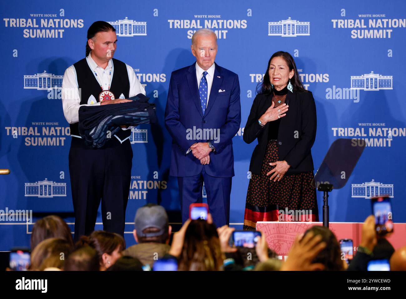 Il Segretario degli interni degli Stati Uniti Deb Haaland (R) e l'Assistente Segretario per gli affari indiani Bryan Newland (L) presentano al presidente degli Stati Uniti Joe Biden una coperta di ottava generazione proveniente da un'azienda di proprietà tribale ricamata con "Joe Biden Champion for Indian Country prima delle osservazioni al vertice delle Nazioni tribali della Casa Bianca del 2024 al Dipartimento degli interni di Washington, DC, USA, 09 dicembre 2024. L'amministrazione Biden-Harris ha dato priorità al rafforzamento delle relazioni nazione-nazione, onorando la fiducia e gli obblighi del trattato con le tribù riconosciute federalmente, e avanzando la sovranità tribale Foto Stock
