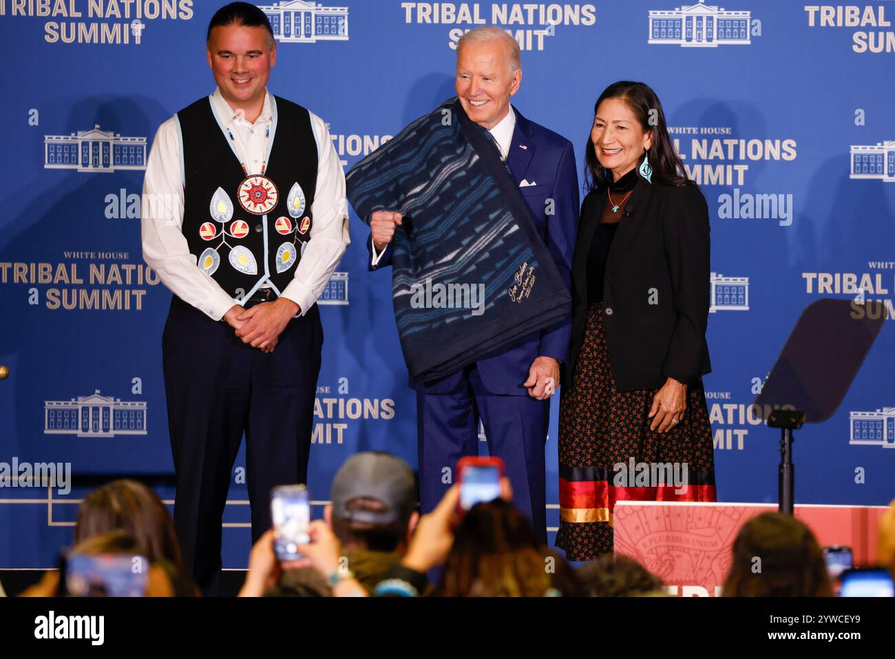 Il Segretario degli interni degli Stati Uniti Deb Haaland (R) e l'Assistente Segretario per gli affari indiani Bryan Newland (L) presentano al presidente degli Stati Uniti Joe Biden una coperta di ottava generazione proveniente da un'azienda di proprietà tribale ricamata con "Joe Biden Champion for Indian Country prima delle osservazioni al vertice delle Nazioni tribali della Casa Bianca del 2024 al Dipartimento degli interni di Washington, DC, USA, 09 dicembre 2024. L'amministrazione Biden-Harris ha dato priorità al rafforzamento delle relazioni nazione-nazione, onorando la fiducia e gli obblighi del trattato con le tribù riconosciute federalmente, e avanzando la sovranità tribale Foto Stock