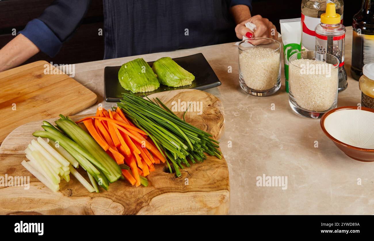 Una tavola di legno splendidamente organizzata con verdure a fette, avocado e riso sushi in bicchieri, preparata per la preparazione del sushi in cucina. Foto Stock