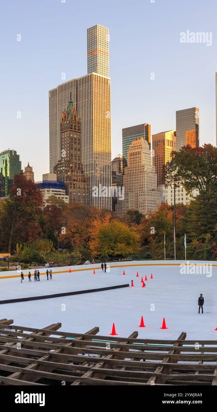 Central Park a New York in autunno. Una pista di pattinaggio su ghiaccio con persone che pattinano e coni rossi è in primo piano, circondata da alberi autunnali. I grattacieli si trainano Foto Stock