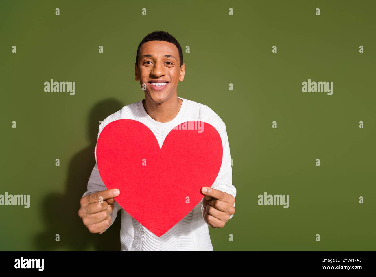 Un giovane con un grande sorriso regge un cuore rosso sullo sfondo cachi, mostrando amore e felicità. Foto Stock