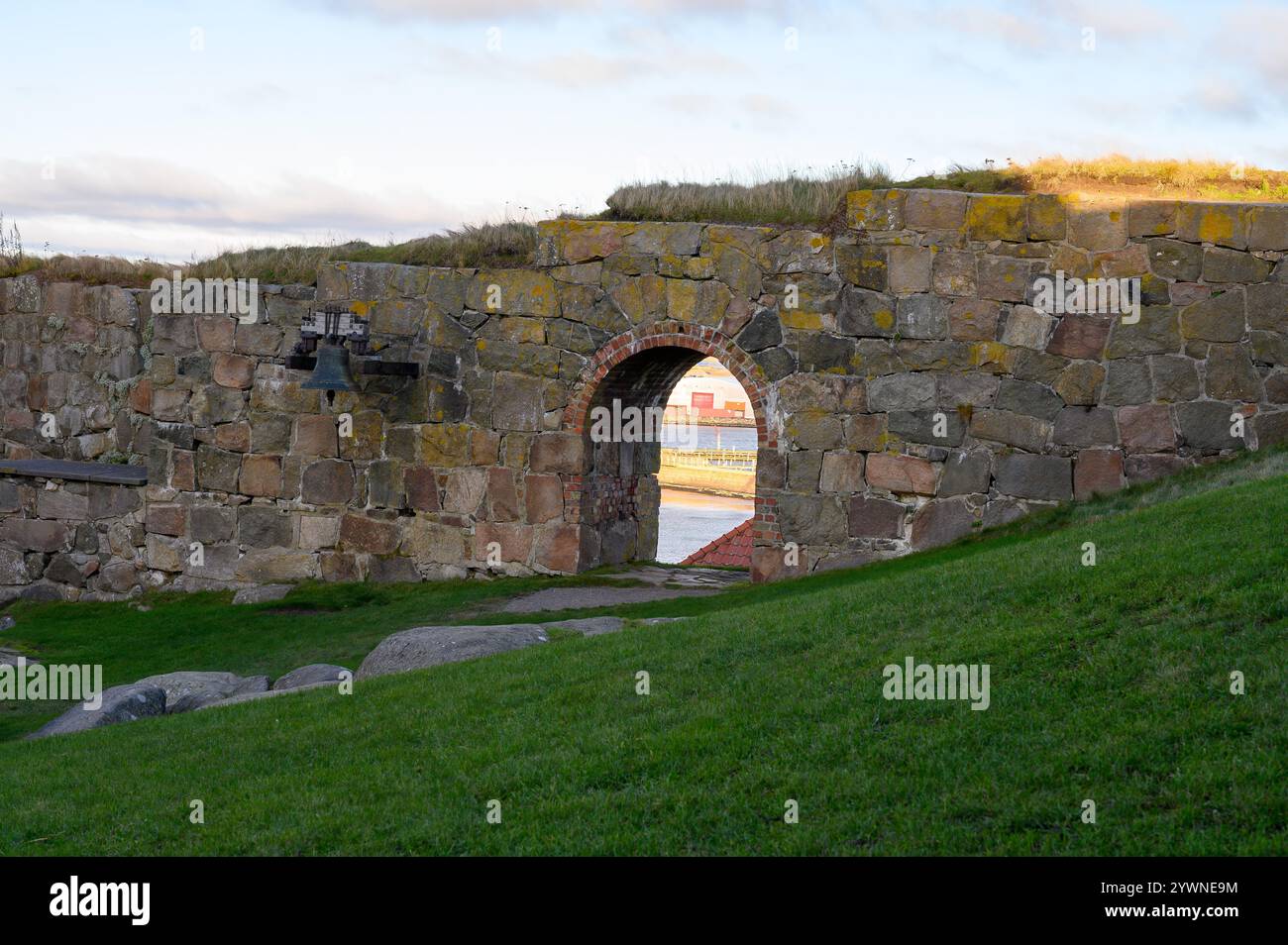 Un muro di pietra con un arco circonda un'area erbosa. La luce del sole diffonde toni caldi nel paesaggio, con un pizzico di nuvole nel cielo blu Foto Stock