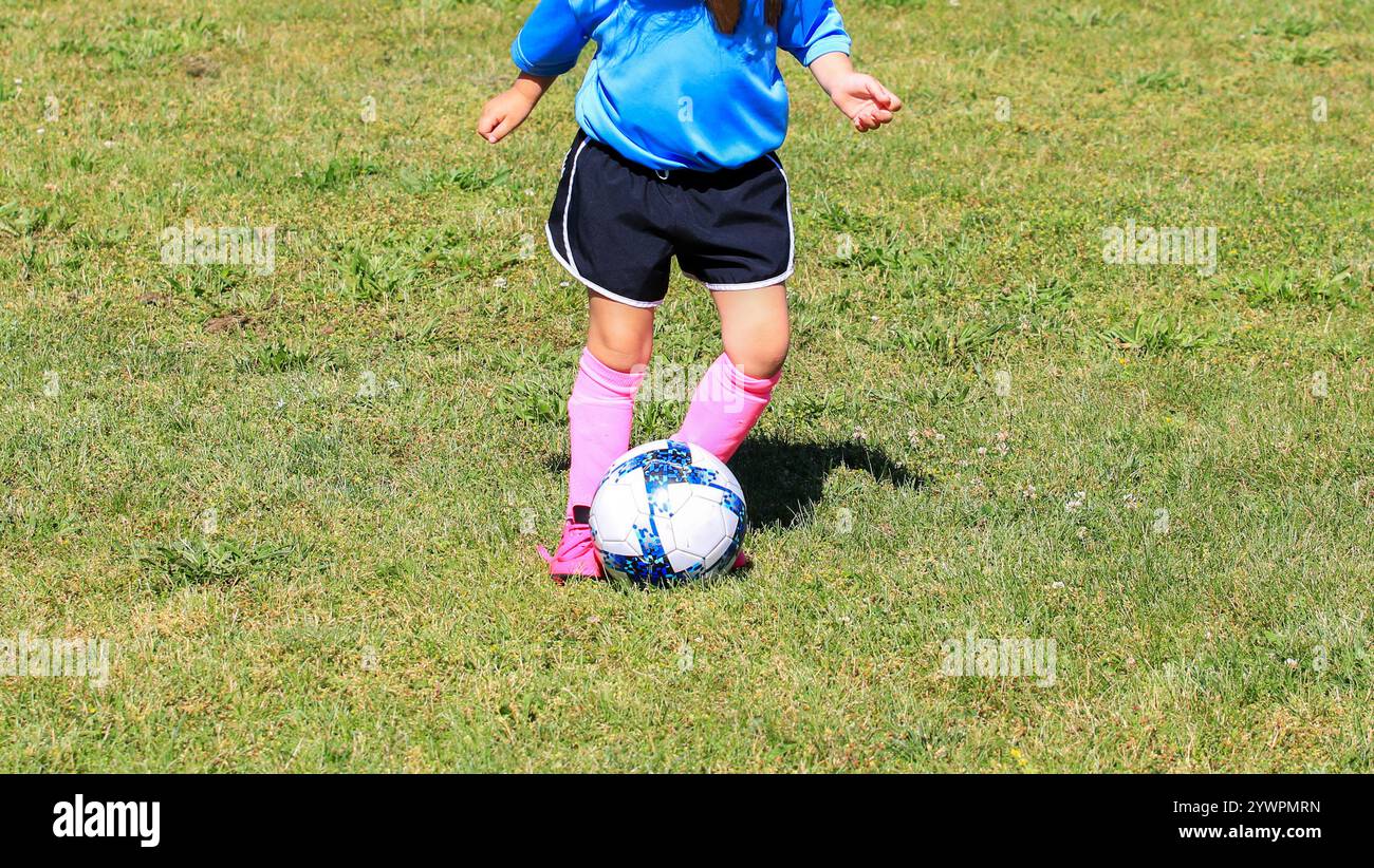 Una ragazza con camicia blu e calze rosa drilla abilmente un pallone da calcio su un campo erboso. Foto Stock
