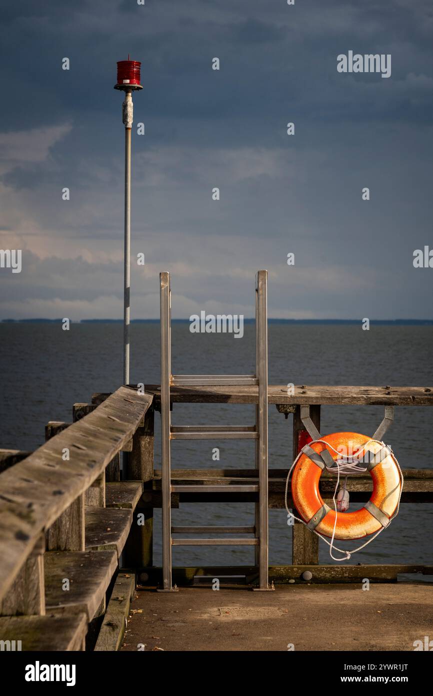 Molo in legno con anello di salvataggio, scala in metallo e luce rossa di sicurezza contro un cielo moody su un mare calmo. Foto Stock