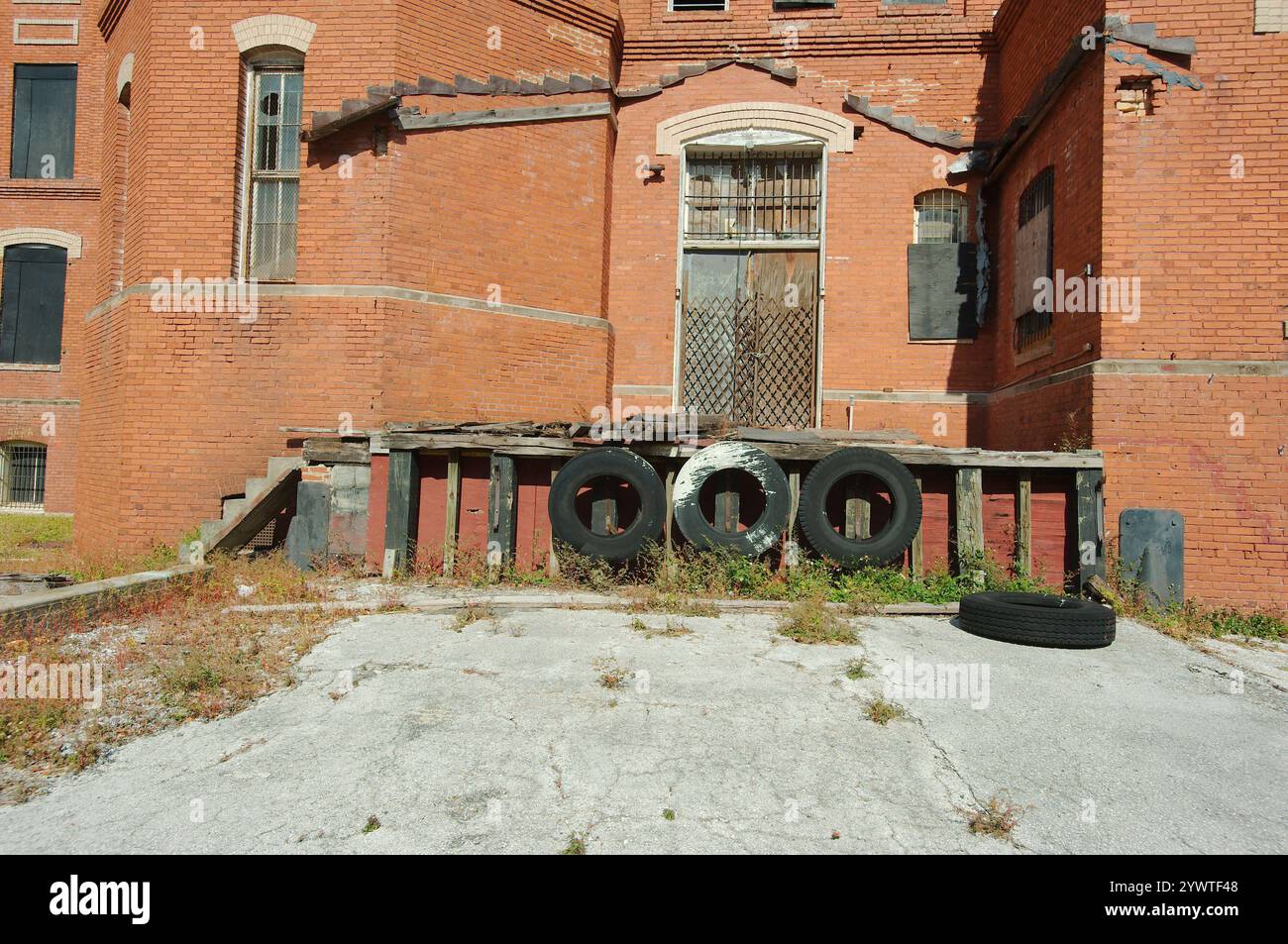 Lato della vecchia parete della banchina di carico in mattoni rossi con tre pneumatici neri sul bordo della banchina. Finestre ad arco coperte nere. Barre di sicurezza in acciaio nero. Foto Stock