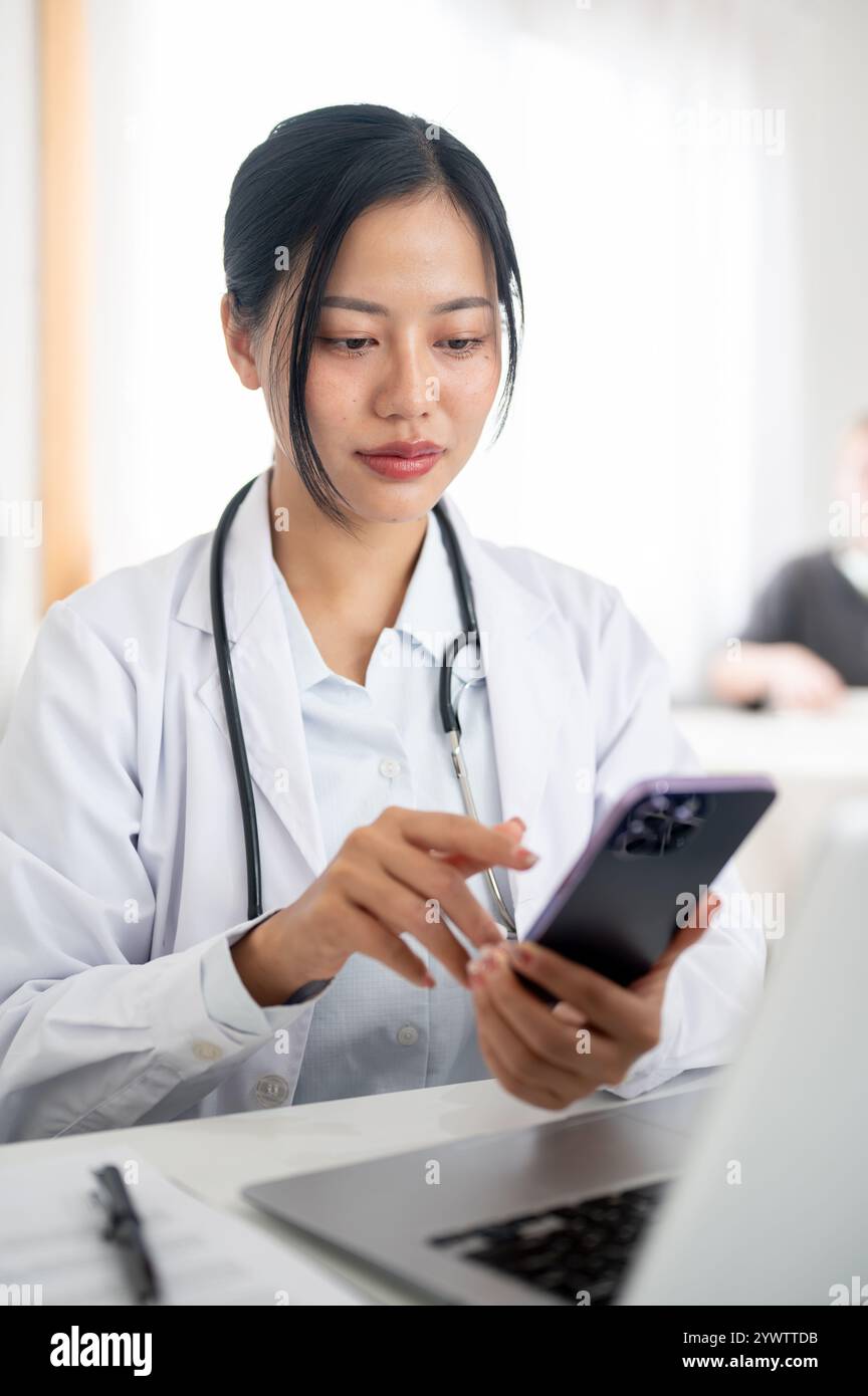 Una bella e positiva dottoressa asiatica in uniforme si siede alla sua scrivania in ufficio, leggendo del testo sul suo smartphone. persone e tecnologia wireless Foto Stock