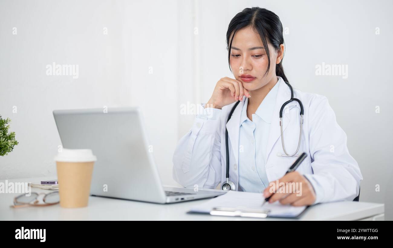 Un'attraente donna asiatica in uniforme, si è concentrata sulla lettura di casi medici su carta e sul suo portatile, lavorando alla sua scrivania in ufficio al Foto Stock