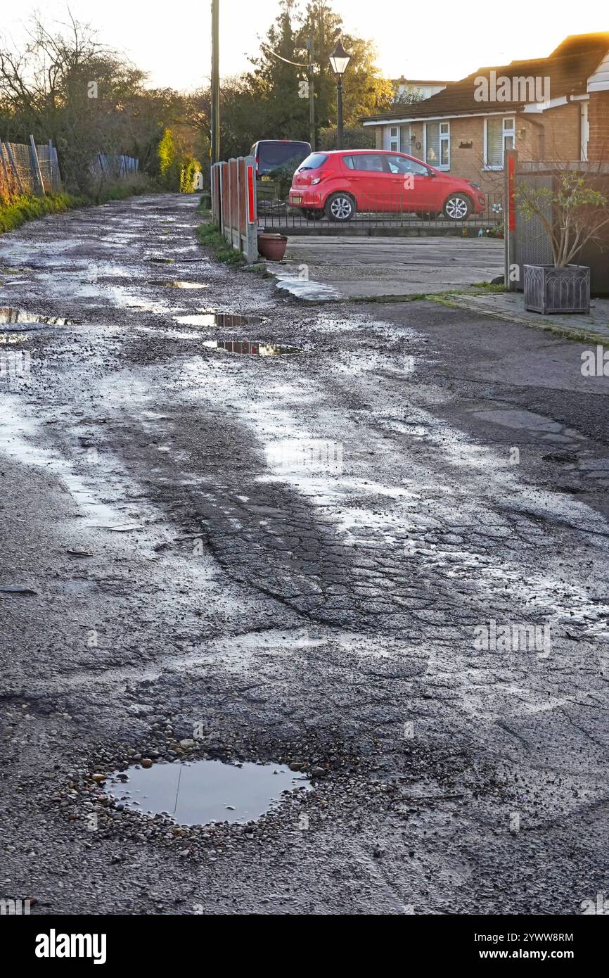 Case accanto non adottate, non fatte, non ricoperte, ricoperte di pozzanghere e bucate, strada bagnata trascurata dopo la pioggia invernale fuori dagli edifici residenziali nel villaggio di Essex nel Regno Unito Foto Stock