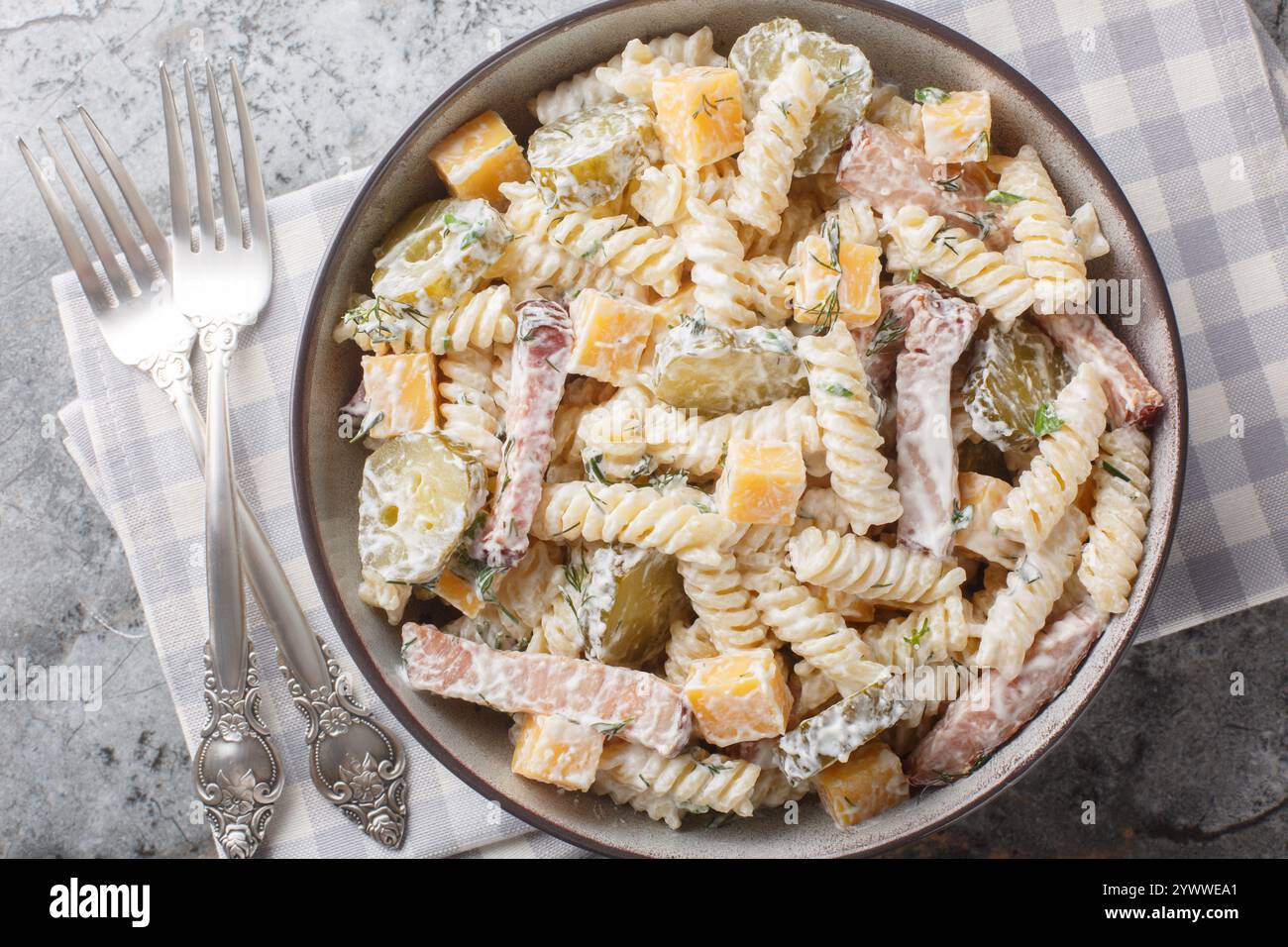 Insalata cremosa di pasta al sottaceti con formaggio giallo, erbe e pancetta in primo piano nel recipiente sul tavolo. Vista dall'alto orizzontale Foto Stock