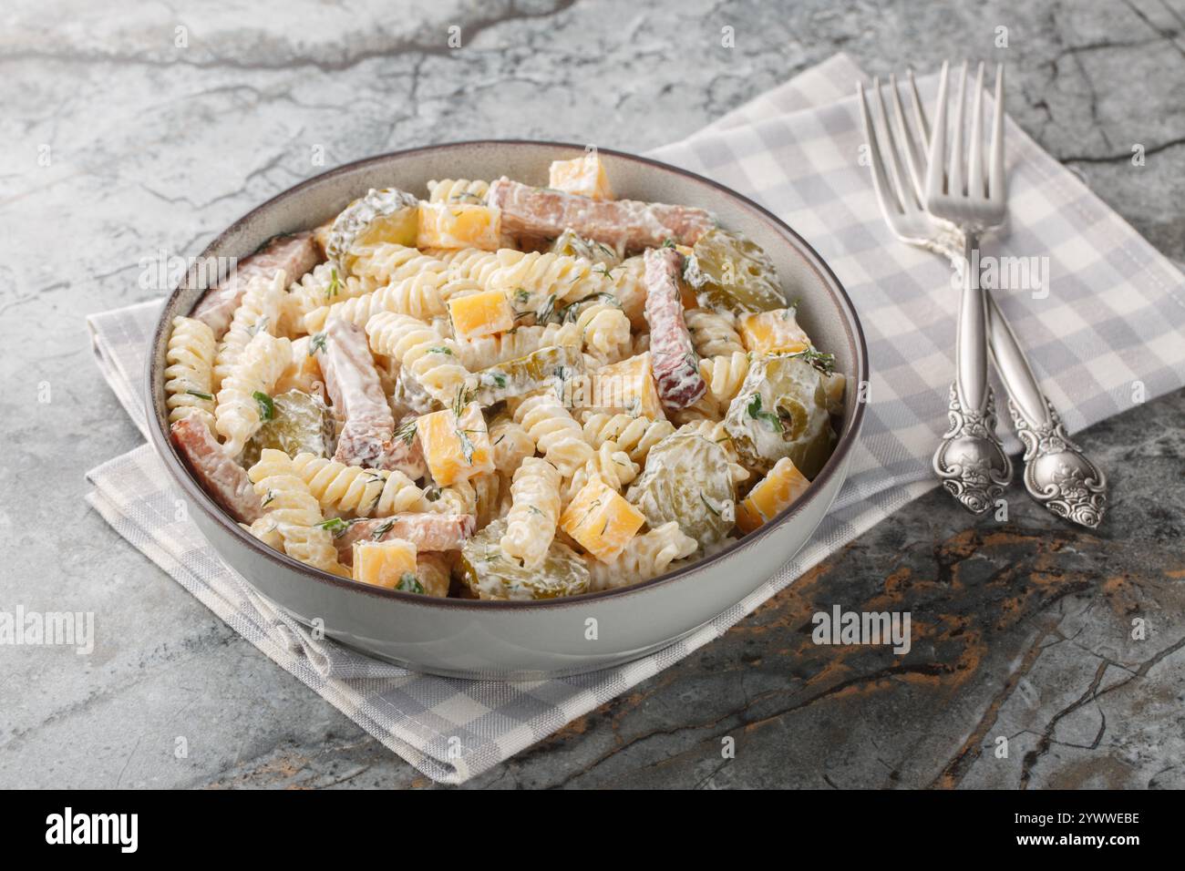 Insalata di pasta invernale deliziosa con sottaceti, formaggio giallo, erbe e pancetta condite con maionese da vicino in una ciotola sul tavolo. Orizzontale Foto Stock