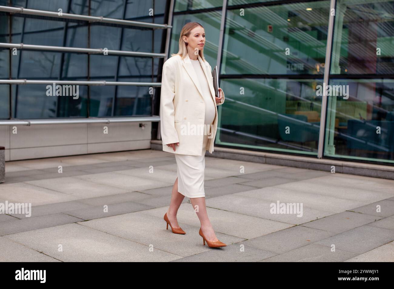 Bella donna bionda incinta con un abito da lavoro bianco con un blazer e un vestito, che cammina all'aperto con un laptop vicino a un edificio aziendale Foto Stock