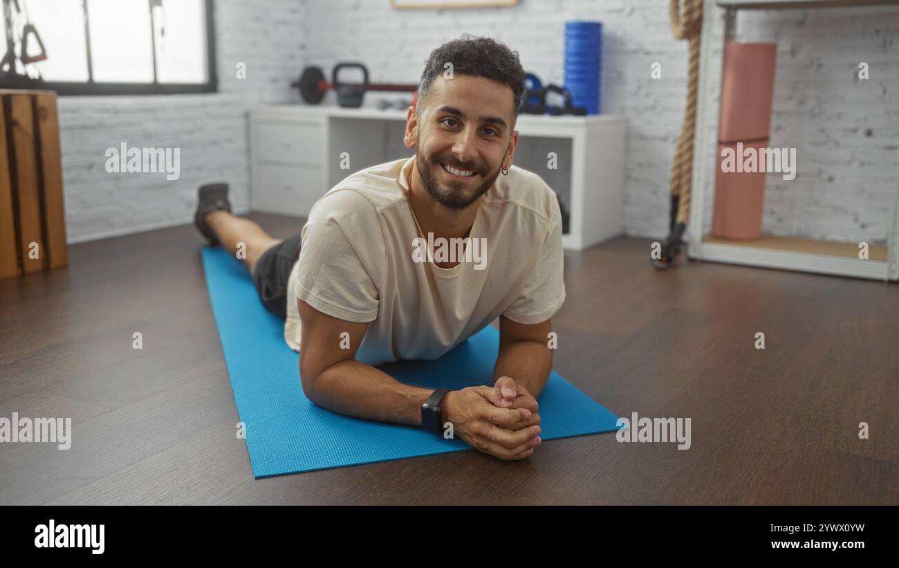 Un bel giovane ispanico giace su un tappetino da yoga sorridendo allegramente in una moderna palestra interna. Foto Stock