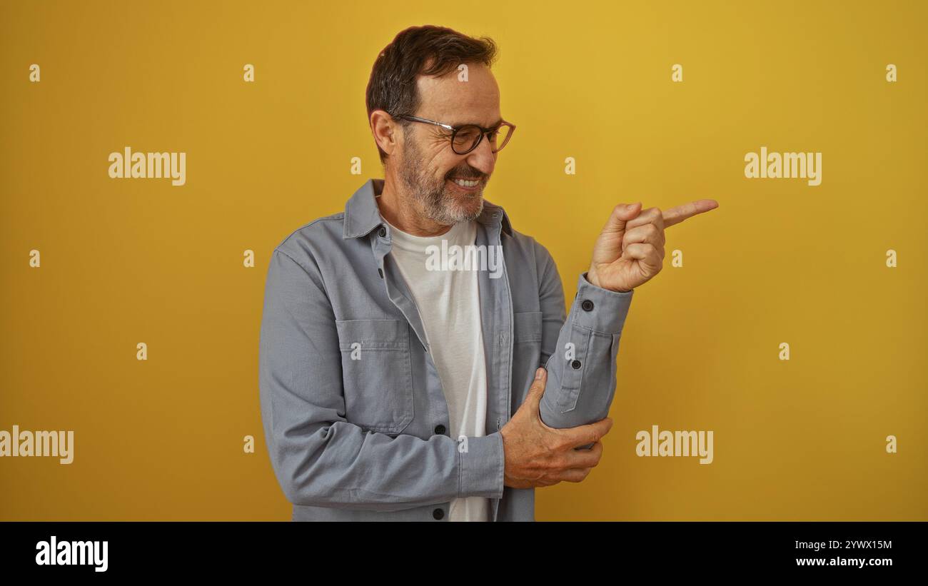 Bellissimo ispanico di mezza età che punta e sorride di fronte a uno sfondo giallo brillante della parete Foto Stock