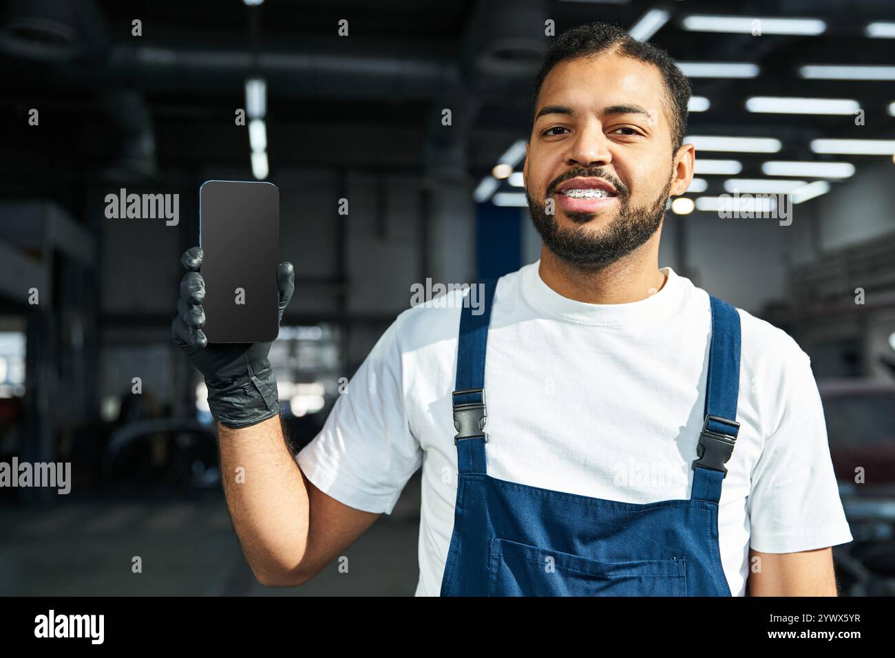 Il giovane meccanico sorride mentre mostra uno smartphone in un ambiente luminoso. Foto Stock