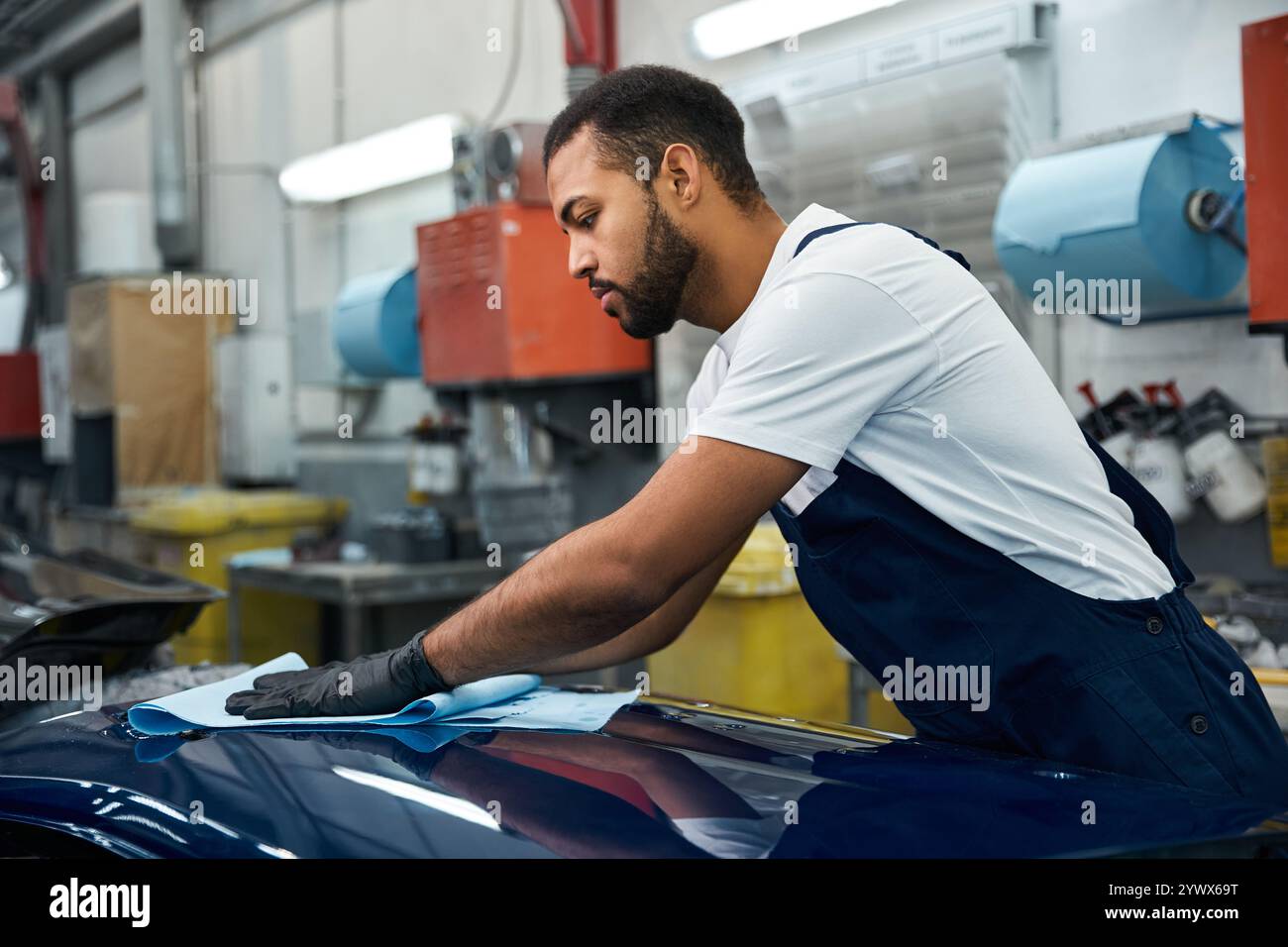 Un giovane meccanico pulisce meticolosamente un veicolo in un'officina di riparazione auto luminosa e affollata. Foto Stock