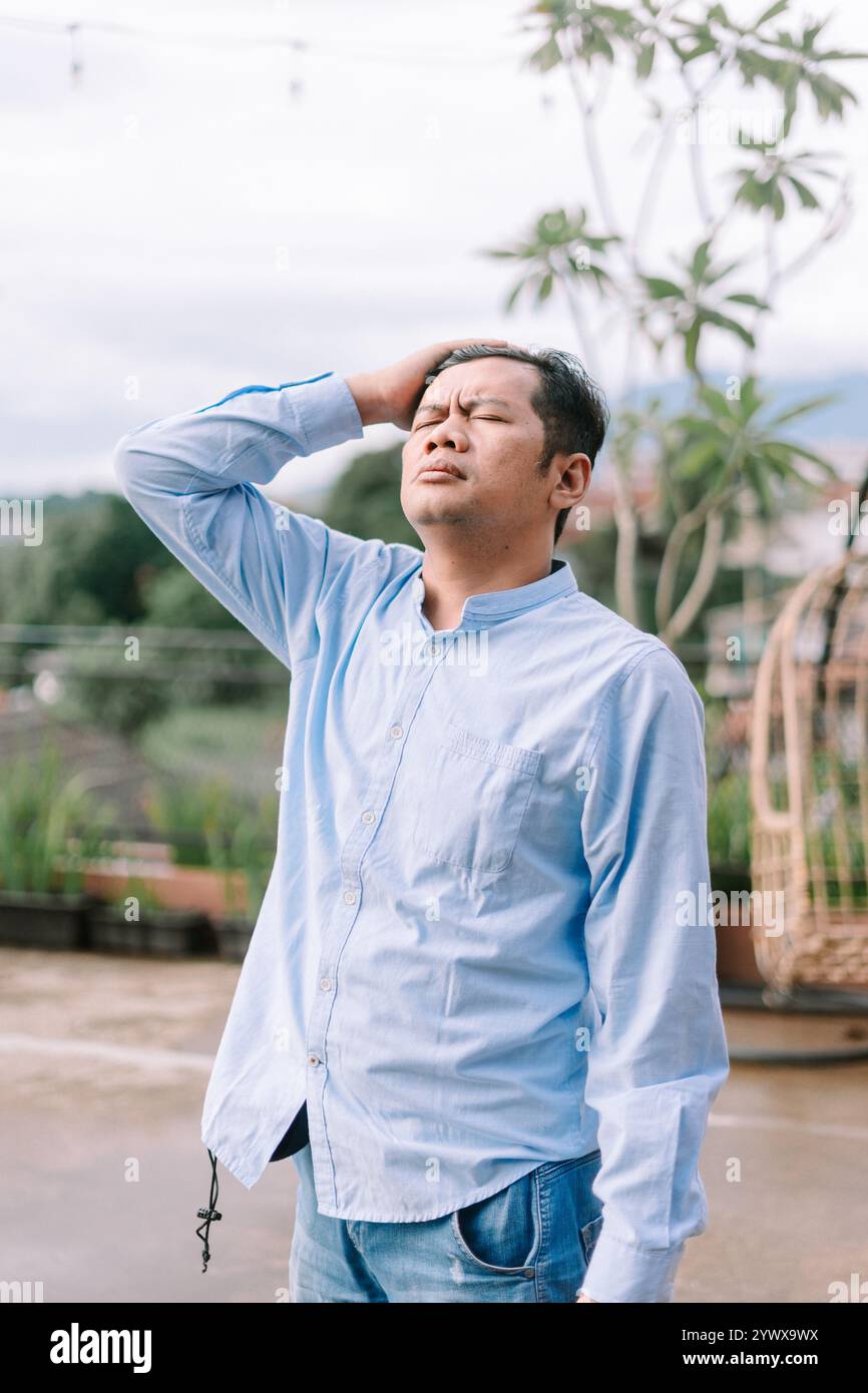 Un uomo del sud-est asiatico che tiene la testa in disagio, avverte visibilmente un mal di testa, cattura un momento di stress fisico o di stress. Foto Stock