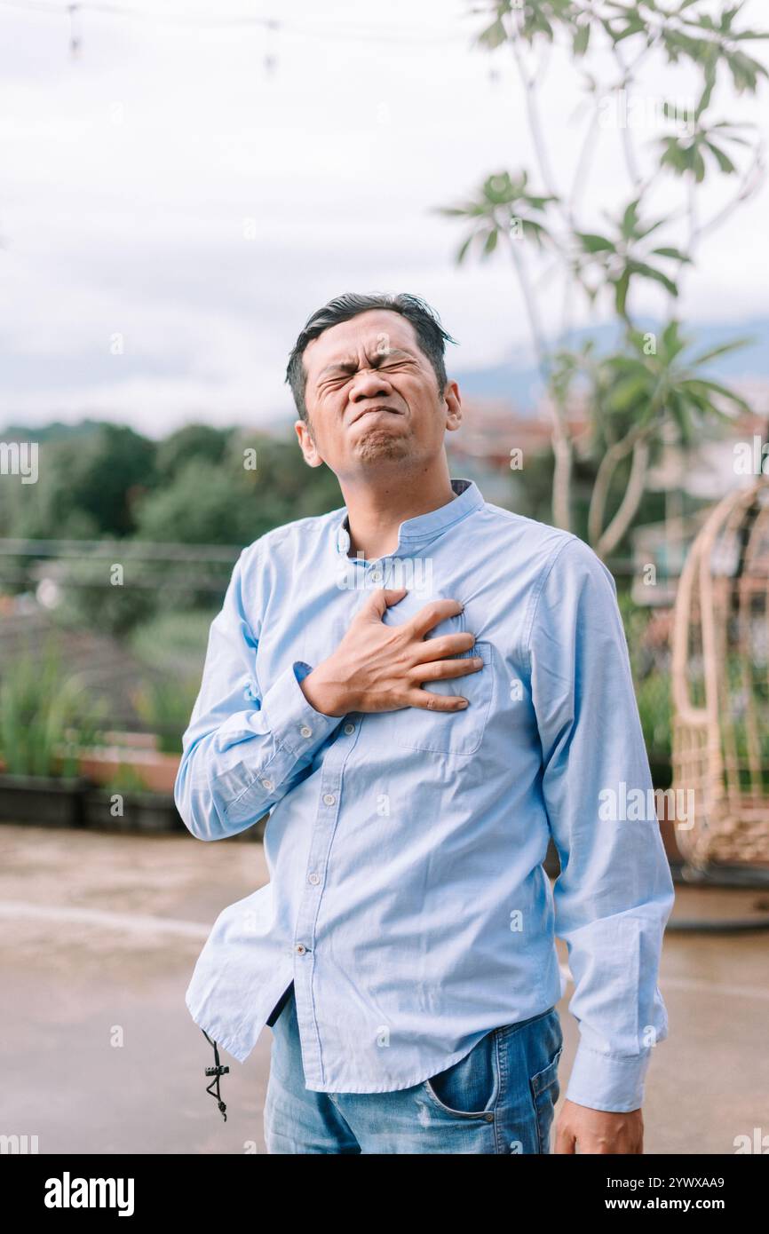 Un uomo del sud-est asiatico che tiene il petto nel dolore, visibilmente afflitto, sperimentando disagio al petto, catturando un momento di preoccupazione per la salute o di medico Foto Stock