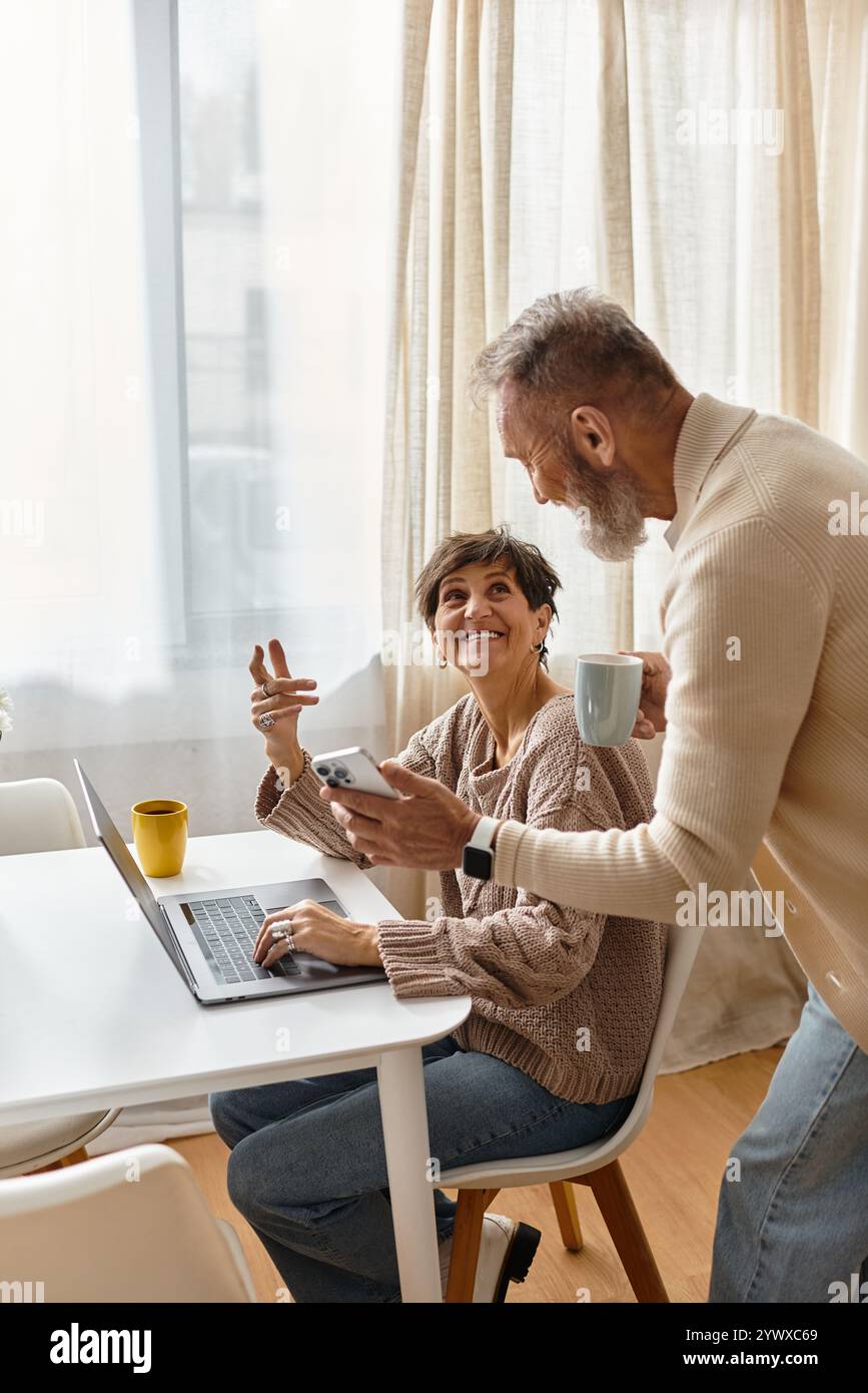 Una donna sorridente lavora sul suo notebook mentre il suo partner la porta con un drink in mano. Foto Stock