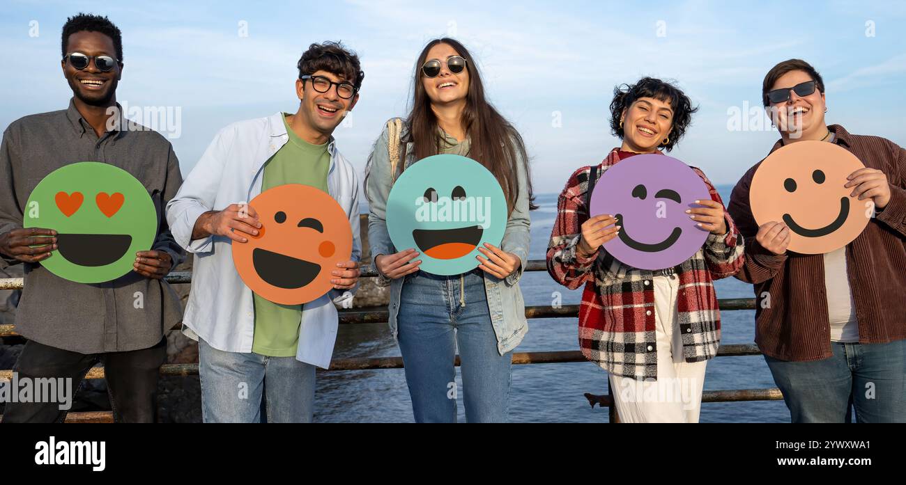 Gruppo eterogeneo di giovani multirazziali con colorate insegne emoji in riva al mare. Amici della Gen Z che esprimono felicità ed emozioni positive attraverso di essa Foto Stock