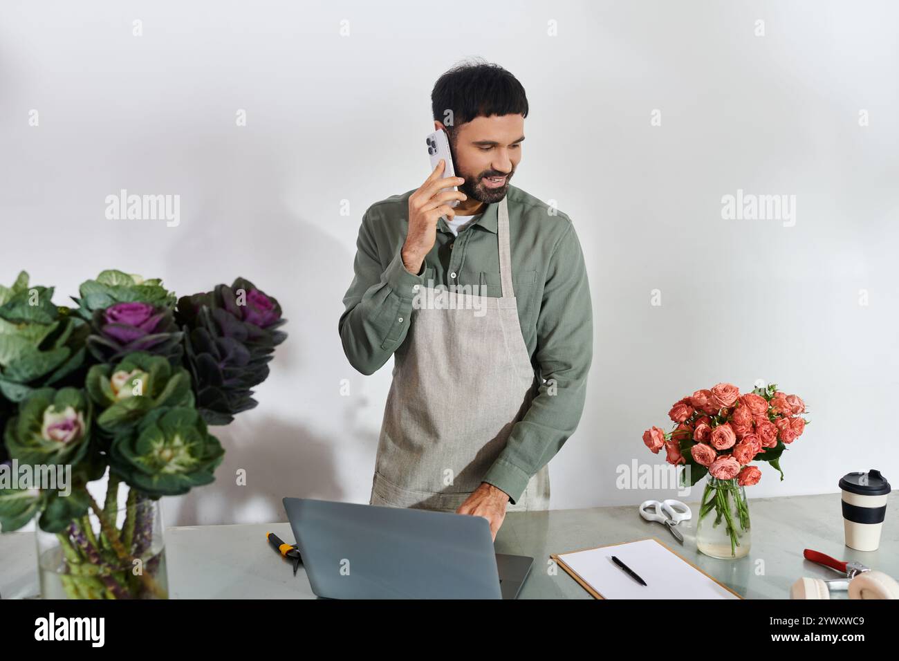 Un bel barbuto parla al telefono mentre organizza fiori freschi nel suo negozio. Foto Stock