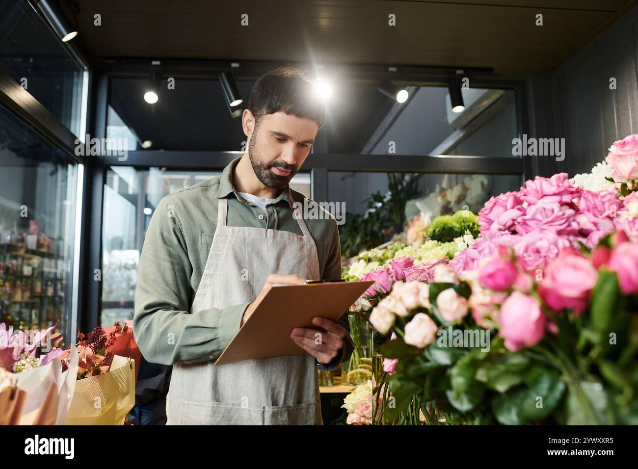 Il bell'uomo barbuto scrive appunti nel suo colorato negozio di fiori, circondato da una flora fiorente. Foto Stock
