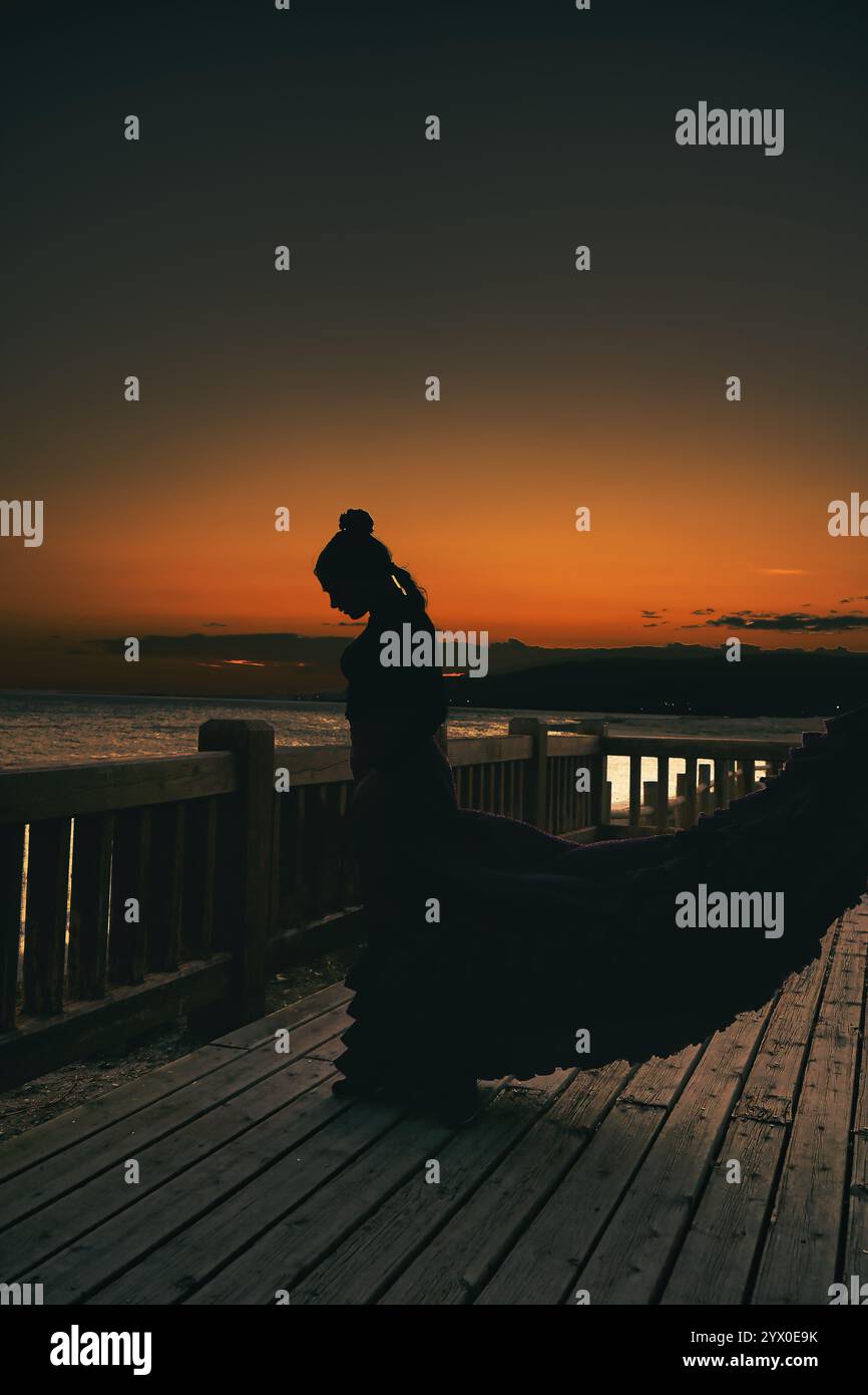 Nella quiete di un tramonto dorato, una silhouette femminile si staglia con grazia contro un cielo che abbellisce di tonalità calde. La donna, avvolta in un'aria di sere Foto Stock