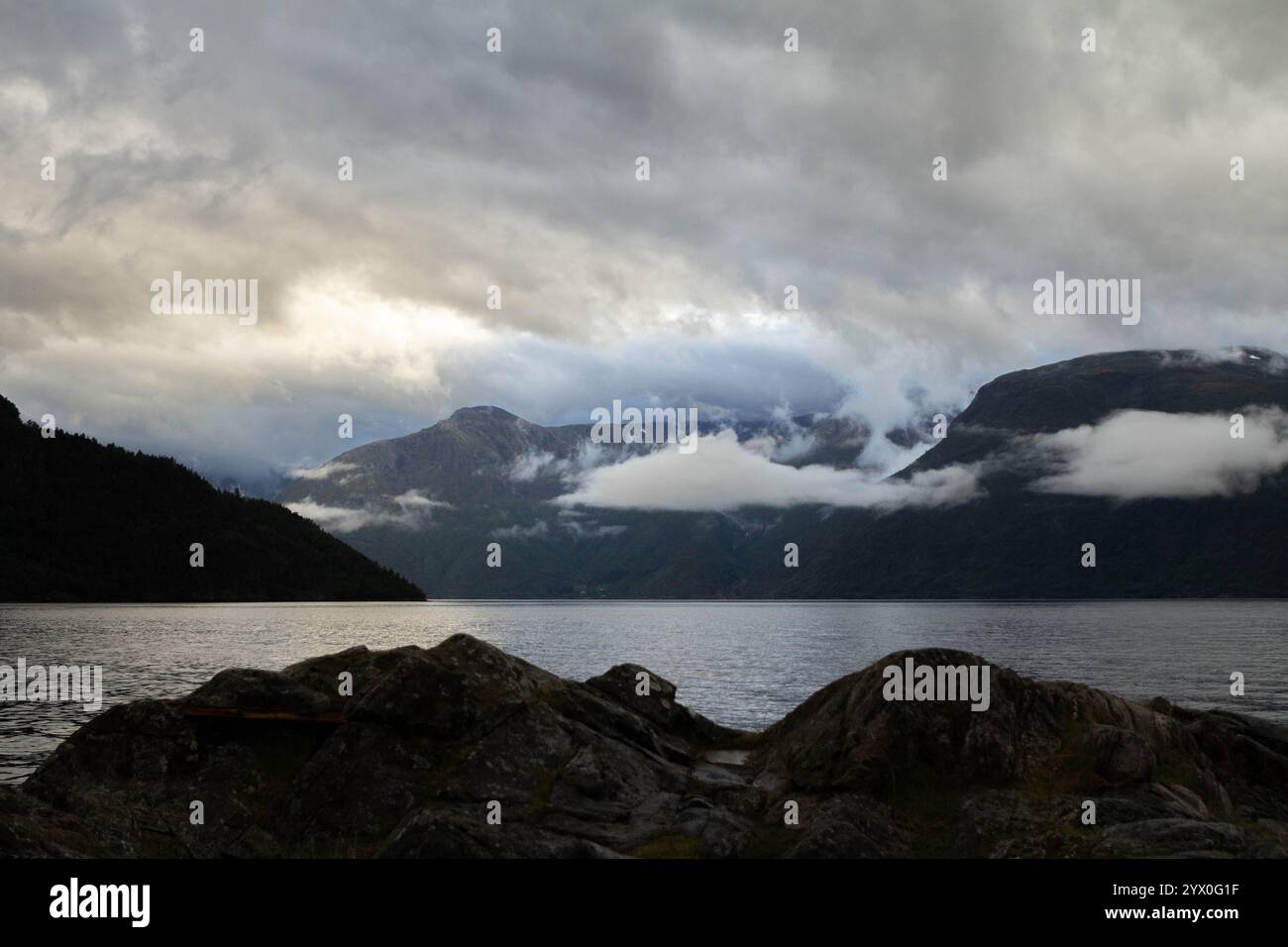 Montagne torreggianti sorgono dalla nebbia, le loro vette frastagliate appena visibili attraverso le nuvole, che si affacciano su un tranquillo fiordo sottostante. Il pettine scenico Foto Stock