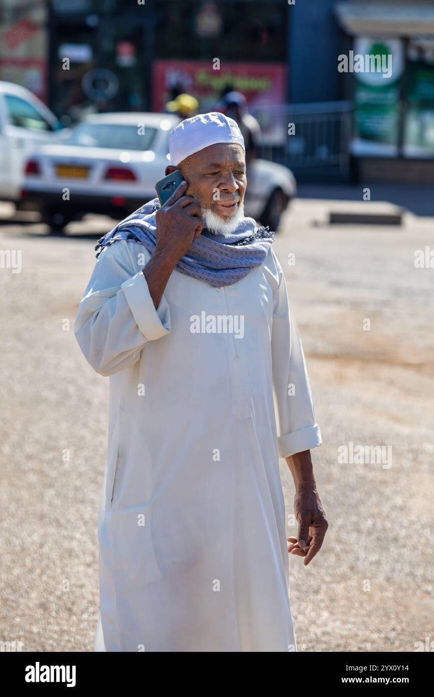 Ritratto di un vecchio musulmano africano che parla sullo smartphone che cammina per strada, con cappello di preghiera taqiyah kufi, uomini Islam all'aperto Foto Stock