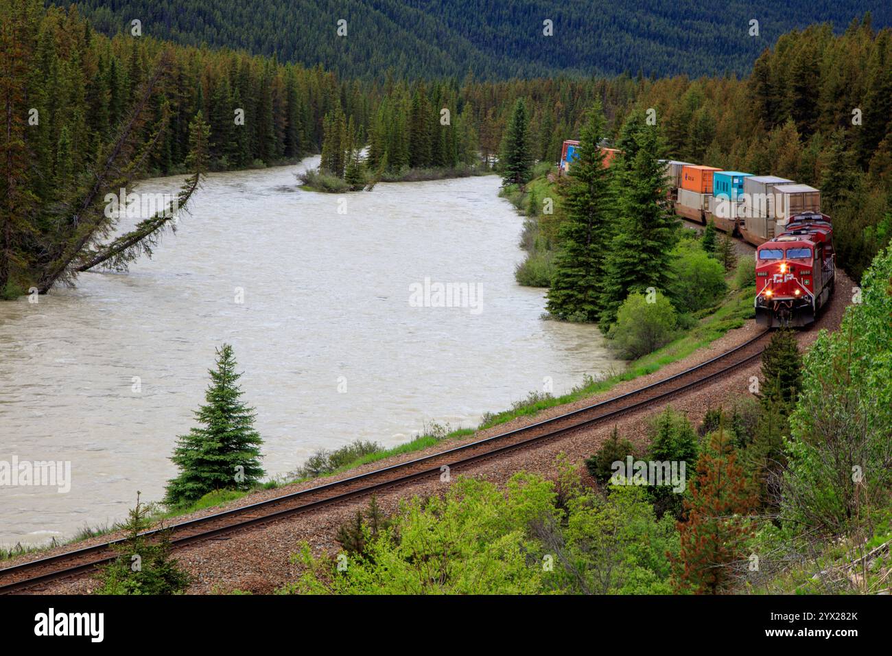 Un treno sta percorrendo un binario vicino a un fiume. Il treno è rosso e trasporta molto carico. Il treno si muove velocemente e l'acqua è a pezzi Foto Stock