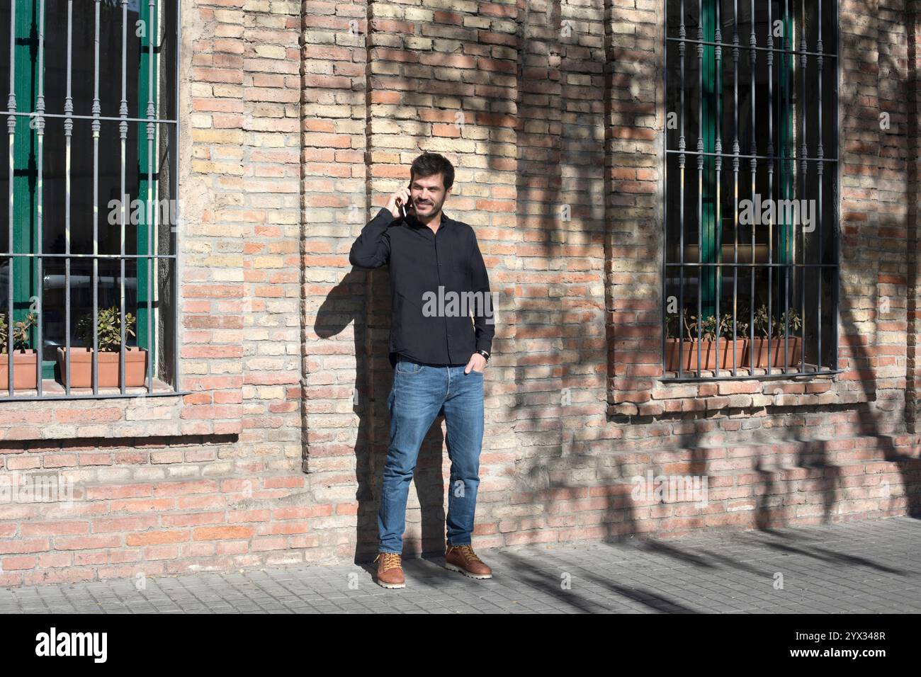 Un uomo caucasico che indossa una camicia nera ride mentre parla al telefono, in piedi contro un muro di mattoni. La luce del sole dona un tocco di gioia. Foto Stock