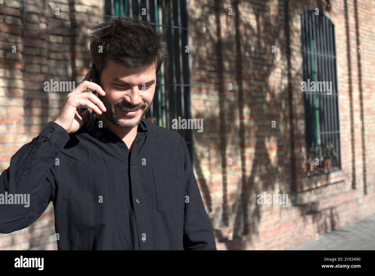 Un uomo caucasico che indossa una camicia nera ride mentre parla al telefono, in piedi contro un muro di mattoni. La luce del sole dona un tocco di gioia. Foto Stock