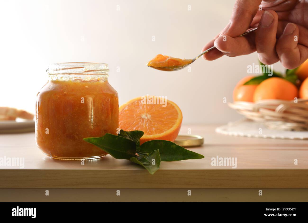 Dettaglio del vaso con marmellata d'arancia e frutta intorno al banco da cucina in legno e mano con un cucchiaio di marmellata e sfondo isolato. Vista frontale; Foto Stock